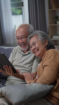 Asian senior couple smiling while video calling on digital tablet computer on sofa in living room