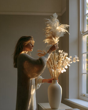 Woman arranging dried flowers in a white ceramic vase by a window