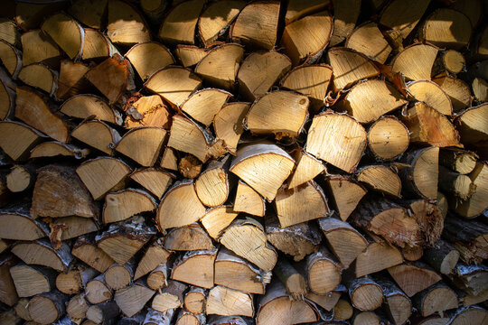 large stack freshly cut firewood logs rough bark warm golden-brown wood grain illuminated sunlight, creating rustic natural texture.