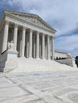 United States Supreme Court Building in Washington, DC. The monumental Neoclassical Revival facade features a grand portico with Corinthian columns beneath a dramatic cloudy blue sky.