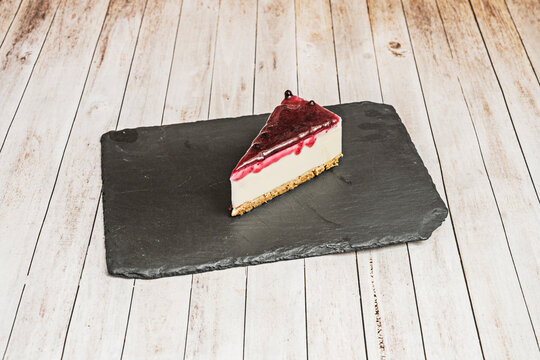 Creamy no-bake cheesecake slice with glossy blueberry compote topping dripping over white filling and graham cracker crust, on a rustic slate board over pale wooden planks
