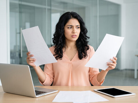 indian woman comparing business documents with a confused expression in a modern corporate office