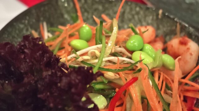 Close-up of mixed vegetable salad with bright green edamame, shredded carrot, thin cucumber strips, bean sprouts, red leaf lettuce, and chili slices tossed in light dressing in dark ceramic bowl, mo