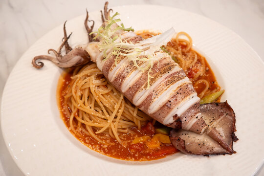Grilled whole squid spaghetti pasta with tomato sauce on white plate.