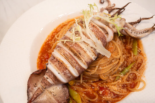 Grilled whole squid spaghetti pasta with tomato sauce on white plate.