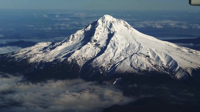 Aerial shot from an airplane window of the large, stunning snow-capped Mount Hood in Oregon, with Cascade Range peaks in the distance under a bright, clear sunny blue sky, flying away from Portland.