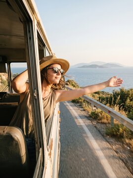 Woman leaning from moving van with arms outstretched on coastal road beside the sea. Road trip and summer travel. Scenic coastal drive and vacation adventure. Freedom and exploration
