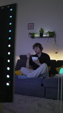 Vertical screen Young man develops an unhealthy habit of eating while watching television at home distracted by entertainment and unaware of how much food he consumes during screen time
