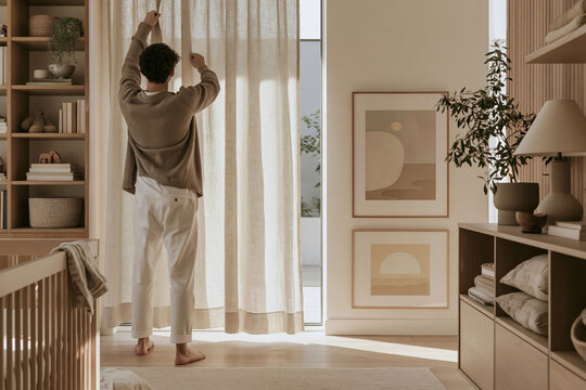 Young man hanging linen curtain panel in softly lit minimalist nursery interior