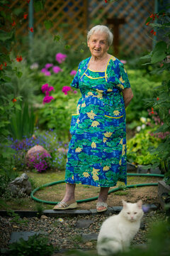 Elderly woman standing in a colorful garden with a white cat blurred in the foreground