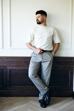 Man with beard posing confidently in stylish casual attire, wearing a cream t-shirt, grey pants, and black loafers