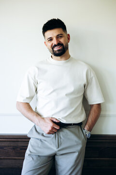 Man with a beard smiling, standing against a wall in a modern room, expressing confidence and a relaxed attitude