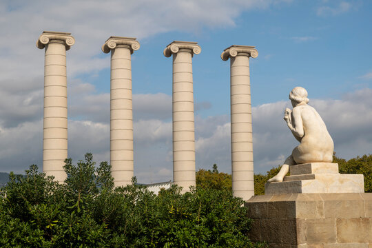 Barcelona, Spain, August 13, 2026 - Four tall Ionic columns stand beside seated stone sculpture on Montjuic hill, framed by green shrubs beneath a cloudy blue sky.