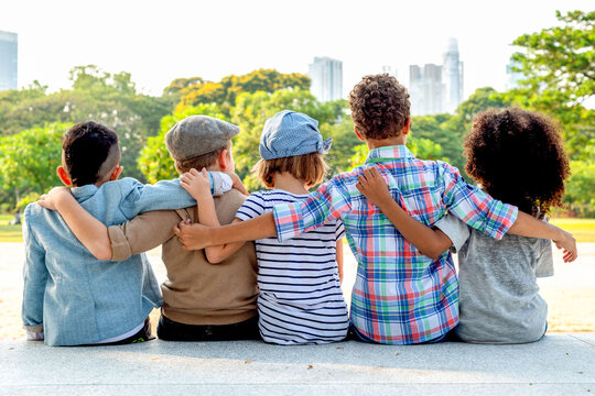 Five children sitting outdoors with arms around each other, backs facing.