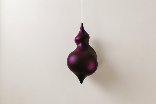 A unique Christmas bulbous, deep purple decorative object hangs suspended by a thin string against a neutral, textured background, casting a soft shadow.