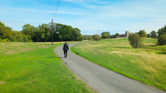 Christian man walking toward rural church along country road. Believer going to church through green countryside. Religious journey to Christian worship service beside fields and trees