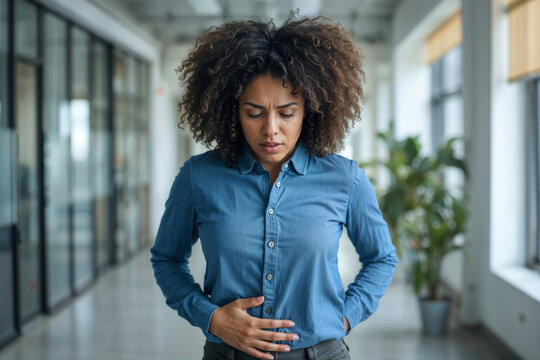 Young black woman stands in office hallway, clutching stomach. She appears uncomfortable, possibly experiencing digestive discomfort