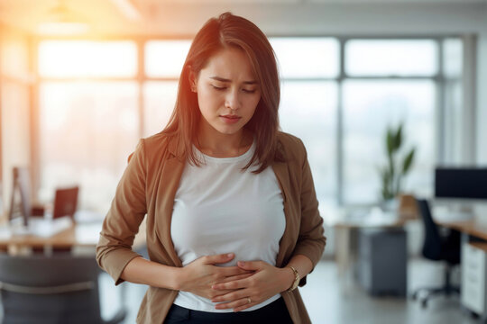 A young Asian woman experiencing stress holds her stomach while working in a bright, modern office
