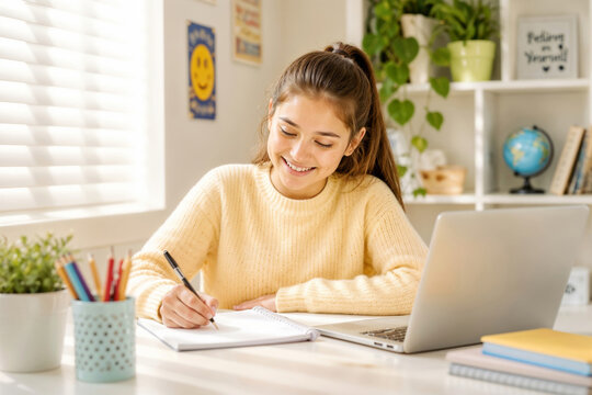 Young girl smiles while writing at desk. Laptop sits beside her notebook. Window lets in soft, natural light. Wall has cheerful smiley poster