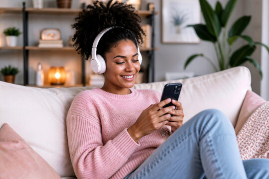 Happy young Afro teenage woman in a pink sweater sits on white couch wearing wireless headphones. Looks at smartphone screen relaxing at home