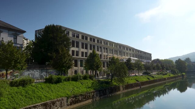 Wide shot of the historic Center Rog creative hub building along the green banks of the Ljubljanica River in Ljubljana.