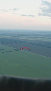 Paratrike glides smoothly over expansive fields under soft sunset sky showcasing pilot and red parachute against green landscape. scene captures serene aerial movement with distant trees