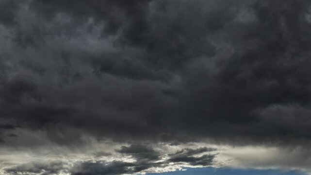 A 4K drone timelapse tilts upward to track dark, ominous storm clouds shifting heavily overhead, completely obscuring the sun while leaving a small sliver of blue sky at the horizon.
