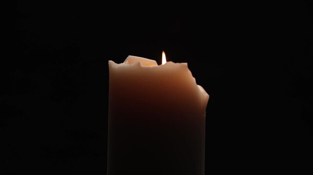 A thick cream-colored candle with an irregular, melted rim, burning with a bright flame and rotates against a black background.