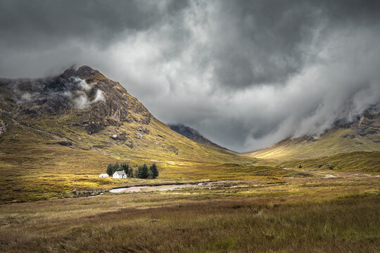 The River Coupall passes by the Wee White House at Glencoe in the Highlands of Scotland with Buachaille Etive Mor shrouded in dark rain clouds behind