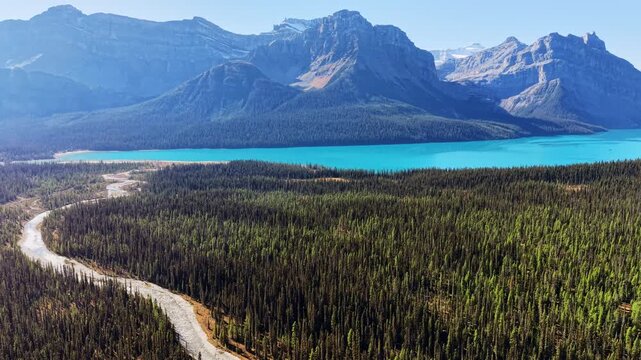 Aerial drone view of a dry gravel riverbed winding through conifer forest toward a turquoise lake and peaks, Banff National Park, Canada.