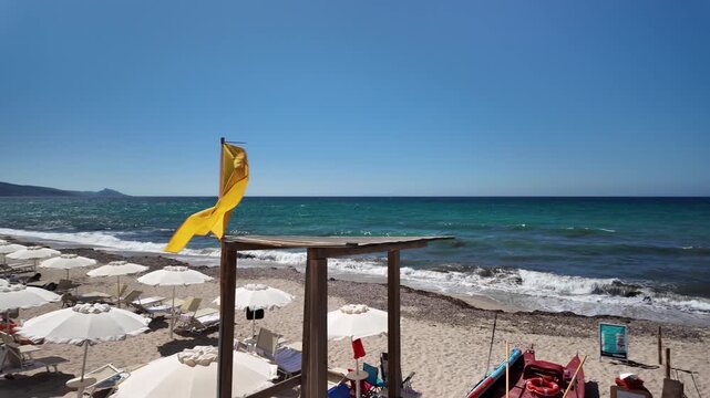 Sunny, sandy beach by turquoise sea featuring rows of white umbrellas and sun loungers wooden lifeguard tower with yellow flag flying, and a mountainous landscape in the background under cloudless sky