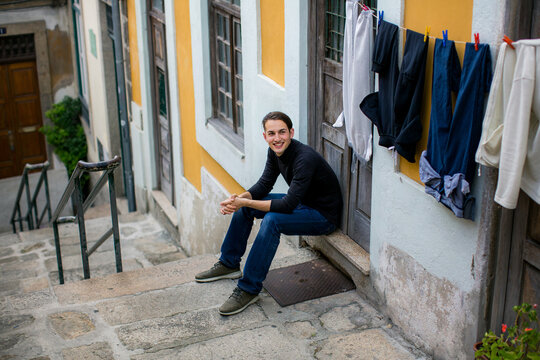 Smiling young man sitting on outdoor steps beside a colorful house with laundry hanging on a clothesline