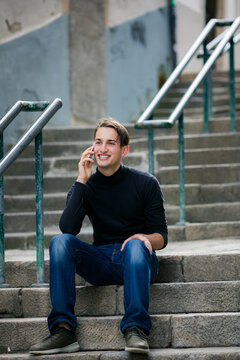 Smiling young man sitting on outdoor concrete steps and talking on a smartphone in an urban setting