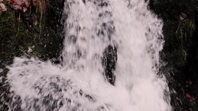 View of multi-tier waterfall flowing over dark moss-covered rock face in deciduous woodland during autumn, silky white water streams contrasted with wet black stone, fallen leaves
