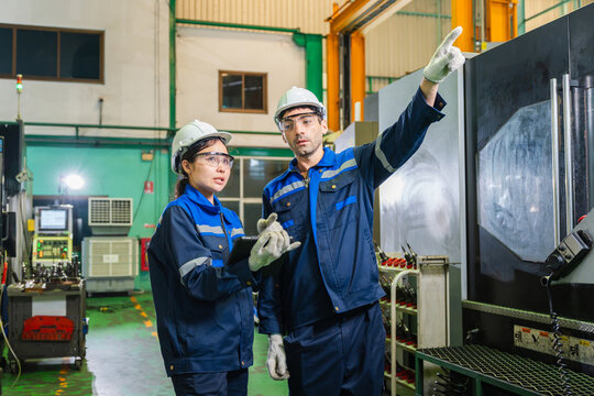 Male and female industrial engineers inspecting CNC equipment with tablet in manufacturing plant, Factory technicians with safety helmets holding tablet and pointing during CNC machine inspection