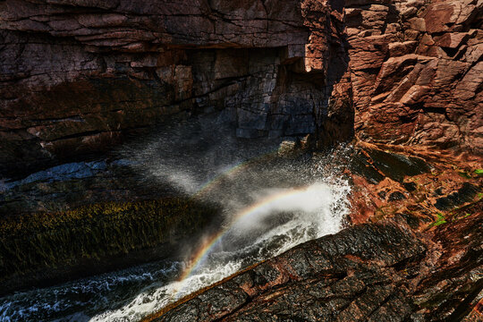  Thunder Hole in Acadia National Park, Maine, featuring a rainbow catching the mist of a crashing wave against the pink granite rock inlet, New England