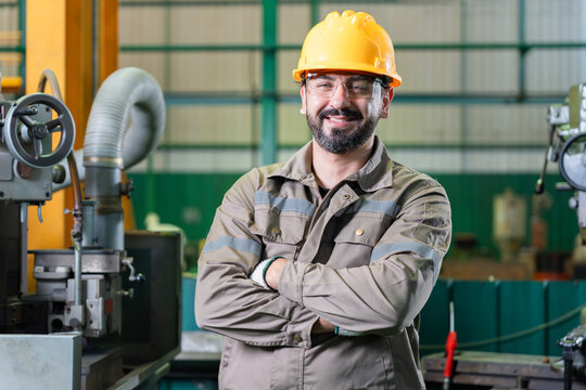 Smiling male industrial engineer standing with arms crossed in manufacturing plant, Confident factory technician wearing yellow hard hat posing in heavy machinery workshop