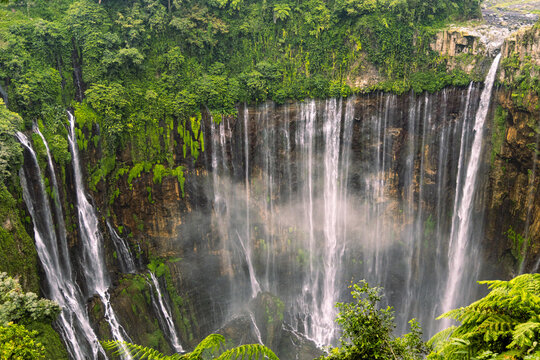 Tumpak Sewu Waterfall in Lumajang, East Java, Indonesia. Hundreds of cascading streams fall from a curved cliff into a lush tropical gorge, surrounded by dense rainforest and rising mist.
