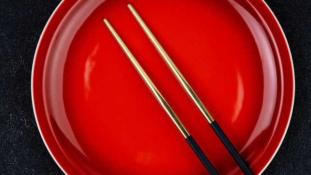 Glossy red plate paired with shiny golden chopsticks arranged neatly in parallel on a dark textured background.