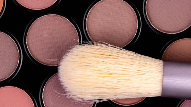 Various shades of eyeshadow arranged in a palette, featuring matte and satin textures in neutral and warm tones. A fluffy makeup brush rests on top, highlighting soft bristles and blending potential.