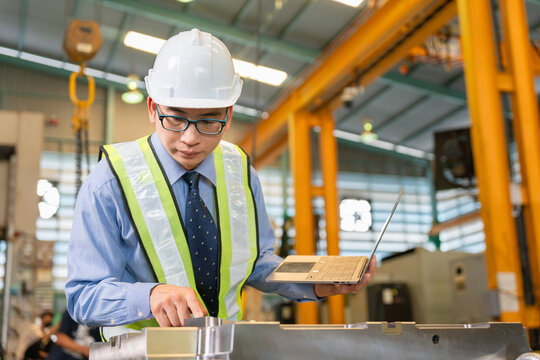 Asian factory manager holding laptop while inspecting metal mold component in manufacturing plant, Industrial engineer using laptop to check metal tooling specs inside heavy machinery workshop