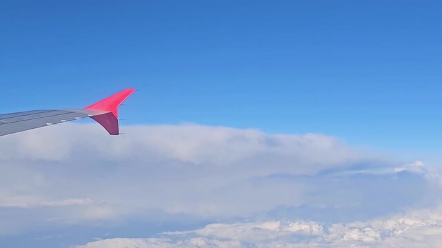 Airplane wing with a red winglet seen through the passenger window during a flight above a thick layer of white clouds on a clear day