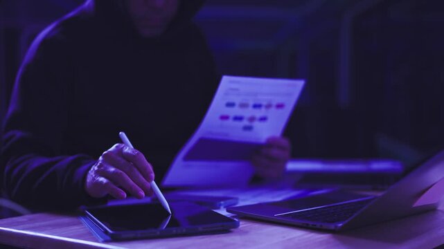 Hacker analyzes flowchart document while touching tablet screen beside laptop in dark room. Cyber espionage threat intelligence, data breach incident investigation, malicious network infiltration.