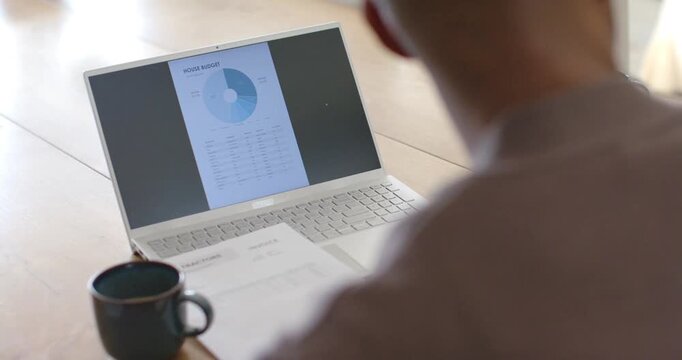 Seated man leaning forward after spotting blue pie chart on laptop while comparing papers at table