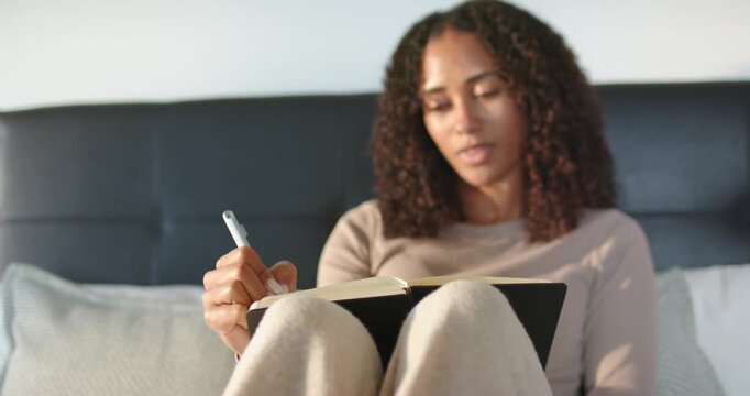 African American woman in 20s balancing black notebook on knees and writing while reflecting on bed