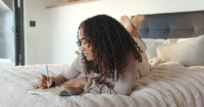 African American woman lying on bed writing in notebook beside smartphone by window, journaling