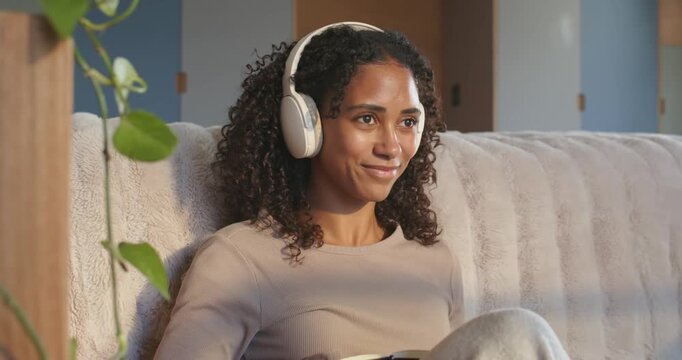 African American woman scrolling phone listening on white headset writing journal with pen on sofa
