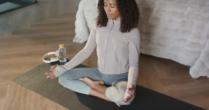 Light urging African American woman meditating on yoga mat in room wearing pink jacket