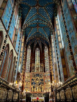 A vertical interior photograph capturing the majestic presbytery of St. Mary's Basilica in Krak&oacute;w with the high Veit Stoss altar, stained glass, and blue starry vaulted ceilings.