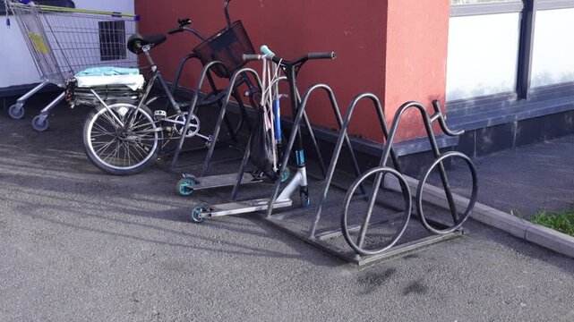 Metal wave-form bike rack anchored beside red-walled building holds bicycle and kick scooter